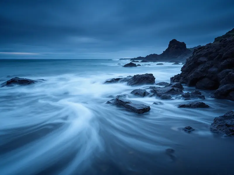 Long-exposure photograph of misty waves on a rocky coast at blue hour